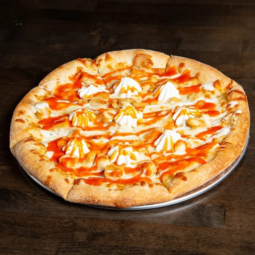 Baba Dough's buffalo chicken pizza, drizzled with hot sauce and white cream, on a dark wooden table.