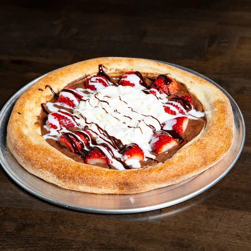 Dessert pizza with chocolate spread, strawberries, whipped cream, and chocolate drizzle on a metal tray.