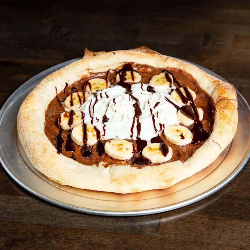 Dessert pizza with Nutella, banana slices, whipped cream, and chocolate drizzle on a metal tray.