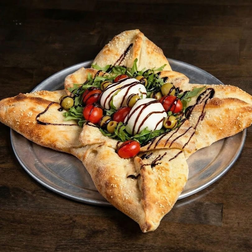 Star-shaped pizza with burrata, cherry tomatoes, olives, arugula, and balsamic glaze on a metal tray.