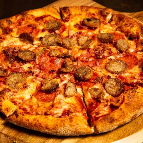 A Baba Dough Pizza with sausage, pepperoni, and bacon on a wooden board.