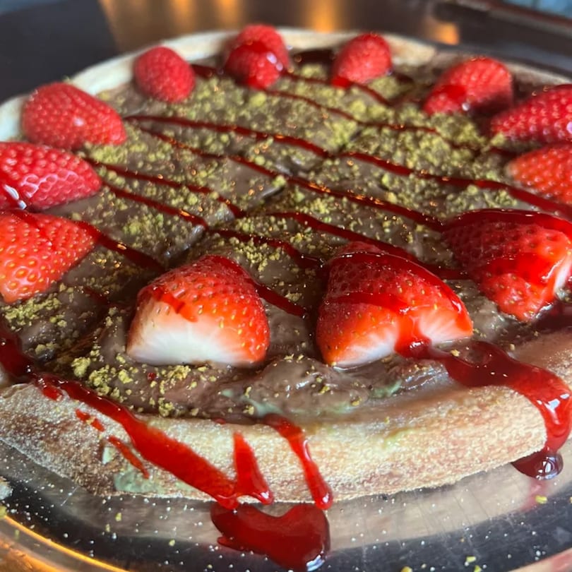 Dessert pizza with chocolate spread, strawberries, pistachio crumbles, and raspberry drizzle.